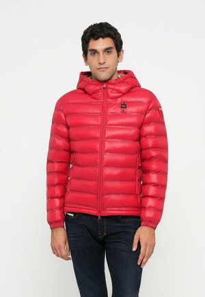 Uomo che indossa una giacca imbottita con cappuccio rossa brillante con zip frontale e jeans scuri, in piedi davanti a uno sfondo bianco uniforme.