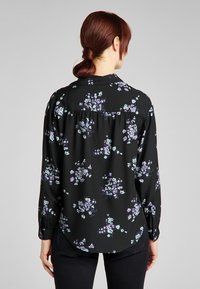 Svart blus med blommönster i lila och blått. Har långa ärmar, krage och en böjd nederdel. Tillverkad av ett glatt, lätt tyg.