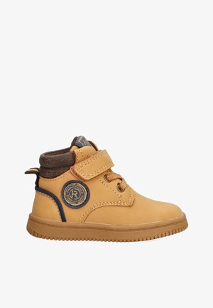 Bota de tobillo de cuero color marrón claro con un acento de tela marrón, correa de velcro y parche de logo circular en el lateral. Suela de goma con agarre texturizado.