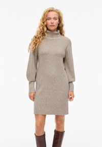 Megztinė suknelė šviesiai smėlio spalvos, su aukštu kaklu, ilgomis pūstomis rankovėmis ir tiesiu kirpimu, su ribbingo audinio tekstūra ir kraštais virš kelio.