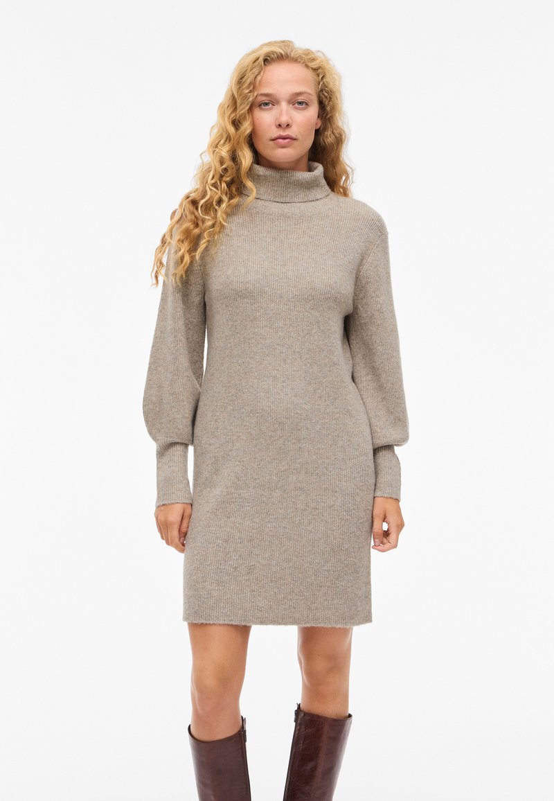 Megztinė suknelė šviesiai smėlio spalvos, su aukštu kaklu, ilgomis pūstomis rankovėmis ir tiesiu kirpimu, su ribbingo audinio tekstūra ir kraštais virš kelio.