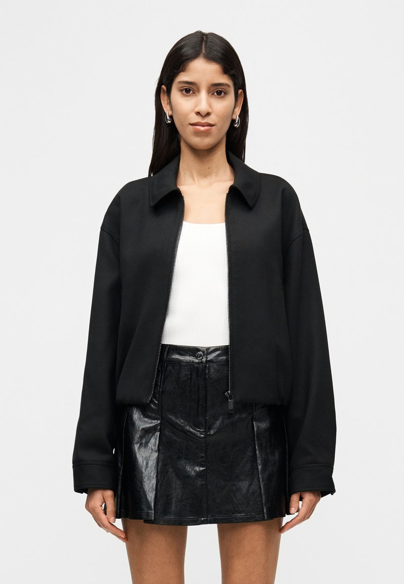 Μαύρο μπουφάν με φερμουάρ, γιακά, φαρδιά γραμμή και μακριά μανίκια, συνδυασμένο με εφαρμοστό λευκό τοπ και λαμπερή μαύρη μίνι φούστα.