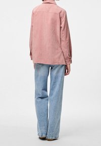 Persoon in een roze geruite jas en lichtblauwe wijde jeans, staande met de rug naar de camera tegen een effen achtergrond.