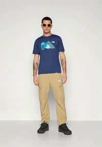 Camiseta azul marino con gráfico de montaña, pantalones cargo beiges, botas de senderismo negras y gafas de sol. Presenta acentos de logo y materiales duraderos.