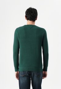 Pull en tricot vert avec un motif texturé, manches longues et col rond, associé à un jean bleu. Ourlet côtelé visible à la taille.