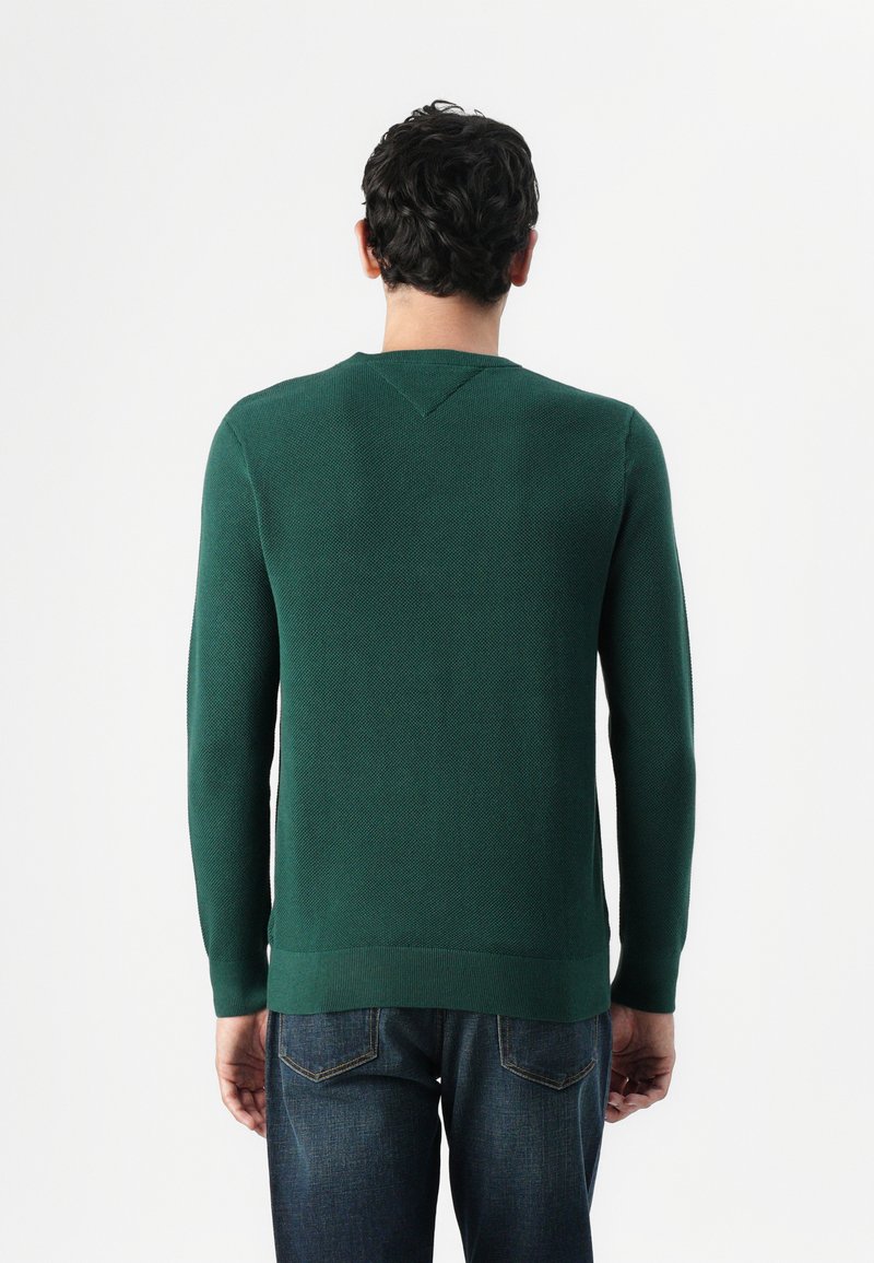 Groene gebreide trui met een textuurpatroon, lange mouwen en een ronde hals, gecombineerd met blauwe jeans. Zichtbare geribbelde zoom op de taille.