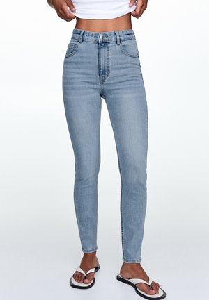 Slim fit light blue denim jeans worn by person lifting white top, paired with white and black flip-flop sandals on plain white background.