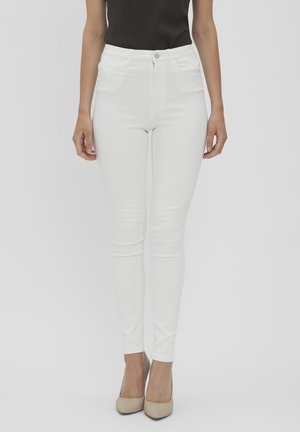 Femme portant un jean blanc taille haute ajusté associé à des talons beiges et un haut noir sans manches sur un fond uni.