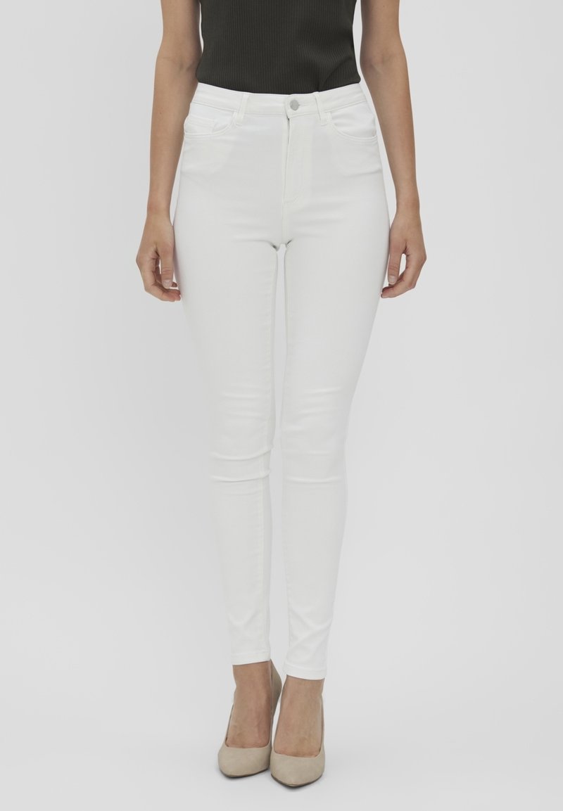 Femme portant un jean blanc taille haute ajusté associé à des talons beiges et un haut noir sans manches sur un fond uni.