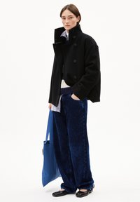 Schwarzer Woll-Doppelreiher-Mantel über einem marineblauen gestreiften Pullover, kombiniert mit weit geschnittenen marineblauen Cordhosen und schwarzen Schuhen, während er eine blaue Tragetasche hält.
