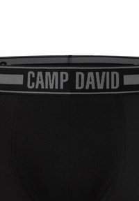 Sort undertøj med en grå elastisk talje, der har "CAMP DAVID" i store bogstaver, glat tekstur, tætsiddende design og ingen synlige mønstre.
