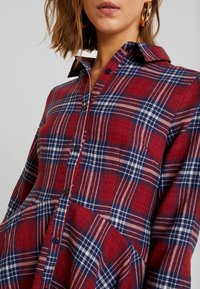 Camicia a quadri rossa e blu con colletto, dotata di bottoni neri e una tasca integrata nel design. Tessuto morbido.