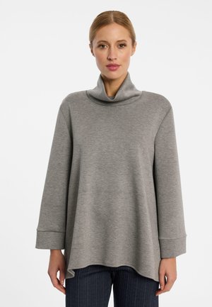 Vrouw draagt een losse grijze longsleeve met hoge kraag en donkere broek, staand tegen een effen witte achtergrond.