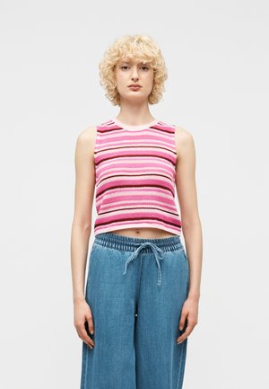 Jeune personne aux cheveux blonds bouclés portant un haut sans manches rayé rose et un pantalon bleu ample à cordon, sur un fond uni.