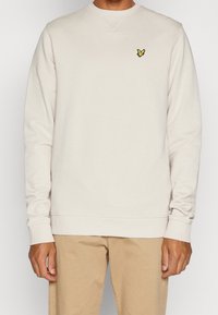 Hombre que lleva un sudadera de manga larga color beige claro con un pequeño pájaro bordado en amarillo y negro en el lado izquierdo del pecho.