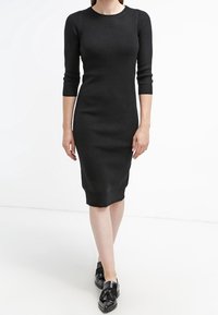 Svart stickad klänning med trekvartsärmar, figurnära silhuett, ribbat halsringning och fåll. Buren med svarta slip-on skor.