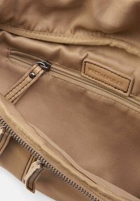 Interno di una borsa di tessuto marrone con una tasca con zip, accessori in argento e un'etichetta in pelle con la scritta "FREDSBRUDER" in rilievo. Tessuto liscio.