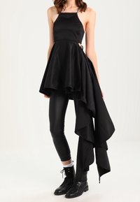 Schwarzes ärmelloses Kleid mit einem ausgestellten Rock, asymmetrischem Saum und einem drapierten Seitendetail, kombiniert mit schwarzen Leggings und Stiefeletten.