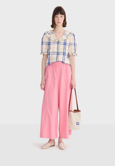 Femme portant un chemisier à carreaux bleu et beige avec un col à volants, un pantalon large rose, des mocassins beiges et tenant un sac fourre-tout beige avec des anses marron.