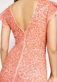 Růžové šaty zdobené flitry s hlubokým výstřihem na zádech, krátkými rukávy a přiléhavým střihem. Obsahují jemné texturované detaily po celé ploše.