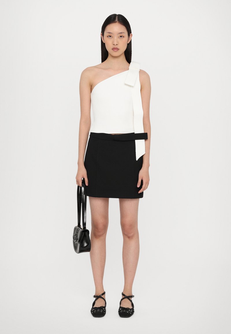 White one-shoulder top, fitted, with a side bow. Black mini skirt with a belt. Black shoes with straps and white accents. Black handbag.