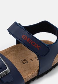 Sandal in pelle blu navy con fibbia argentata, soletta in sughero e logo "GEOX" rosso su una cinghia. Presenta una suola in gomma testurizzata e resistente.
