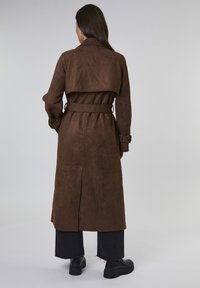 Kobieta stojąca, ubrana w długi brązowy płaszcz z pasem, czarne krótkie spodnie i czarne masywne buty, zwrócona plecami w stronę jednolitego tła.