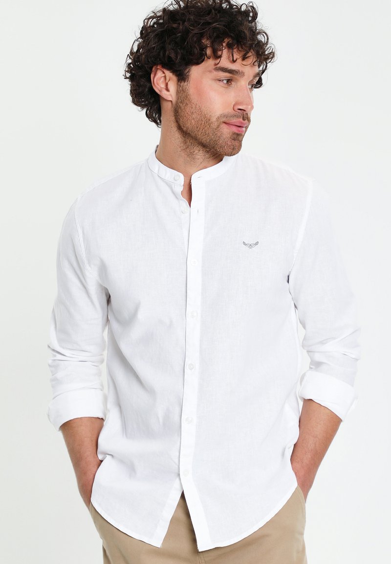 Mann mit lockigem Haar, der ein weißes Hemd mit Stehkragen und hochgekrempelten Ärmeln sowie beige Hosen trägt, die Hände in den Taschen, schaut nach rechts.