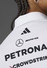 Vit polo skjorta med svarta logotyper: Adidas, Mercedes och AMG, med fet text som läser "PETRONAS" och "CROWDSTRIKE" på framsidan.