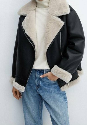 Persona con una chaqueta de cuero negra con forro de borrego beige, un suéter de cuello alto color crema y vaqueros azules, mano en el bolsillo.