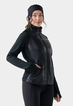 Mujer que lleva una chaqueta negra con cremallera y paneles delanteros acolchados, leggings negros y una diadema negra, de pie con una mano en el bolsillo.