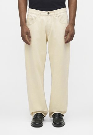 Homme portant un pantalon ample couleur crème et des chaussures en cuir noir sur un fond clair uni, mains relaxées le long du corps.