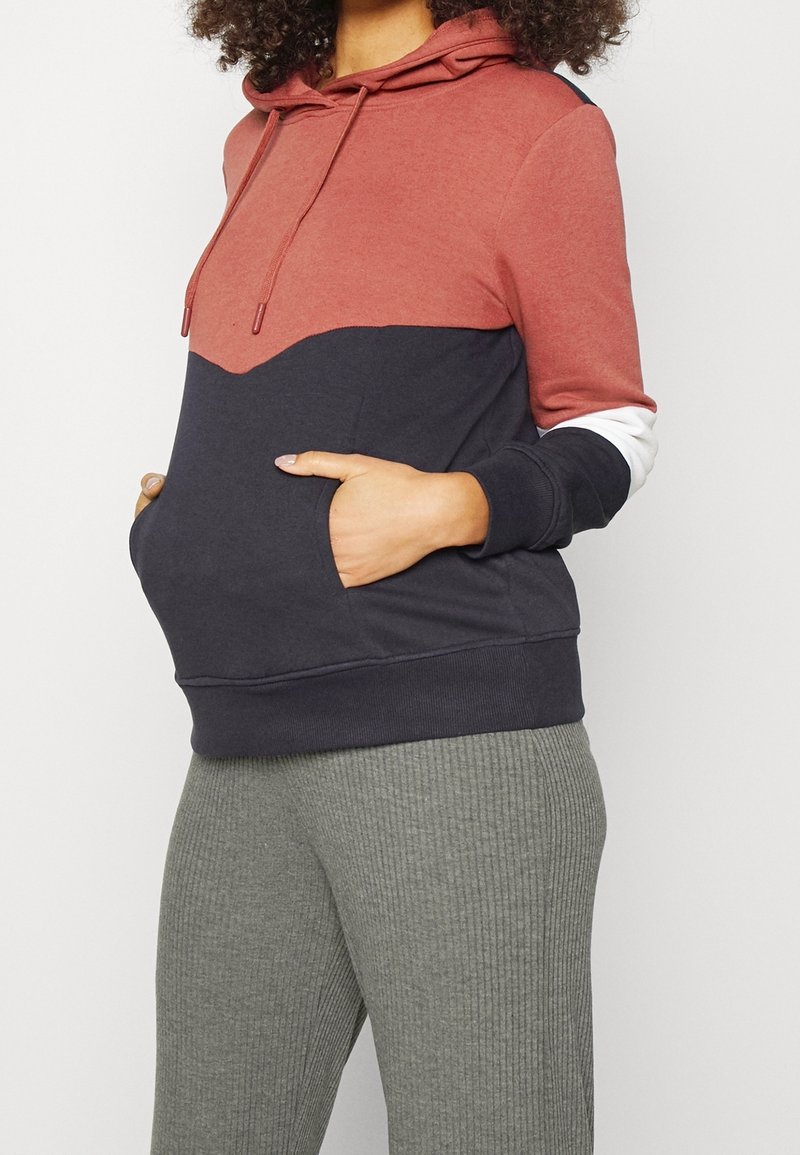 Femme portant un sweat à capuche bicolore rouge et noir avec poches avant et pantalon gris côtelé, mains dans les poches du sweat.
