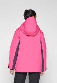 Giacca impermeabile rosa con cappuccio, maniche lunghe e pannelli laterali contrastanti di un violaceo scuro. Il materiale sembra liscio e resistente.