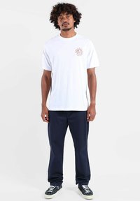 T-shirt blanc en coton à manches courtes, avec un petit logo imprimé en rose. Associé à un pantalon bleu marine et des baskets noires.