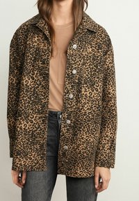 Leopard print jacket in brown tones, featuring a collar, front buttons, and a relaxed fit, paired with a beige top and dark jeans.