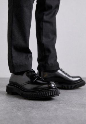 Black leather lace-up shoes with a chunky ridged sole and rounded toe. Worn with tailored dark pants, showcasing polished finish and sleek design.