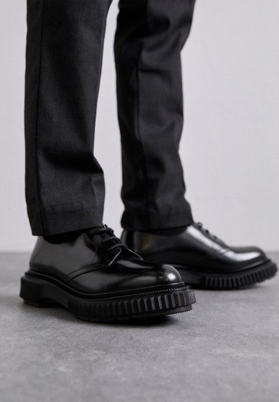 Schwarze Leder-Schnürschuhe mit einer robusten, profilierten Sohle und runder Zehenform. Getragen mit maßgeschneiderten dunklen Hosen, die einen polierten Look und ein elegantes Design zur Schau stellen.