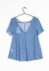 Blusa azul de manga corta con escote en V, fabricada con un tejido ligero y translúcido salpicado de pequeñas estrellas blancas. Cuélga de un gancho negro.