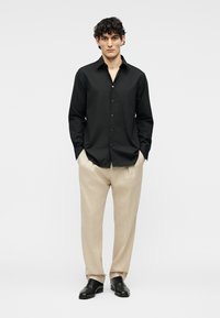 Man with curly hair wearing a black button-up shirt, beige trousers, and black dress shoes, standing with hands in pockets.