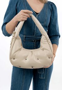 Borsa color crema con una texture liscia, borchie a forma di cuore e un design imbottito. Presenta un manico curvo e dettagli in metallo dorato.