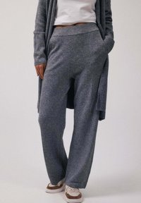 Personne portant un pantalon en maille gris ample et un cardigan assorti par-dessus un top court blanc, debout les mains dans les poches et des baskets blanches.