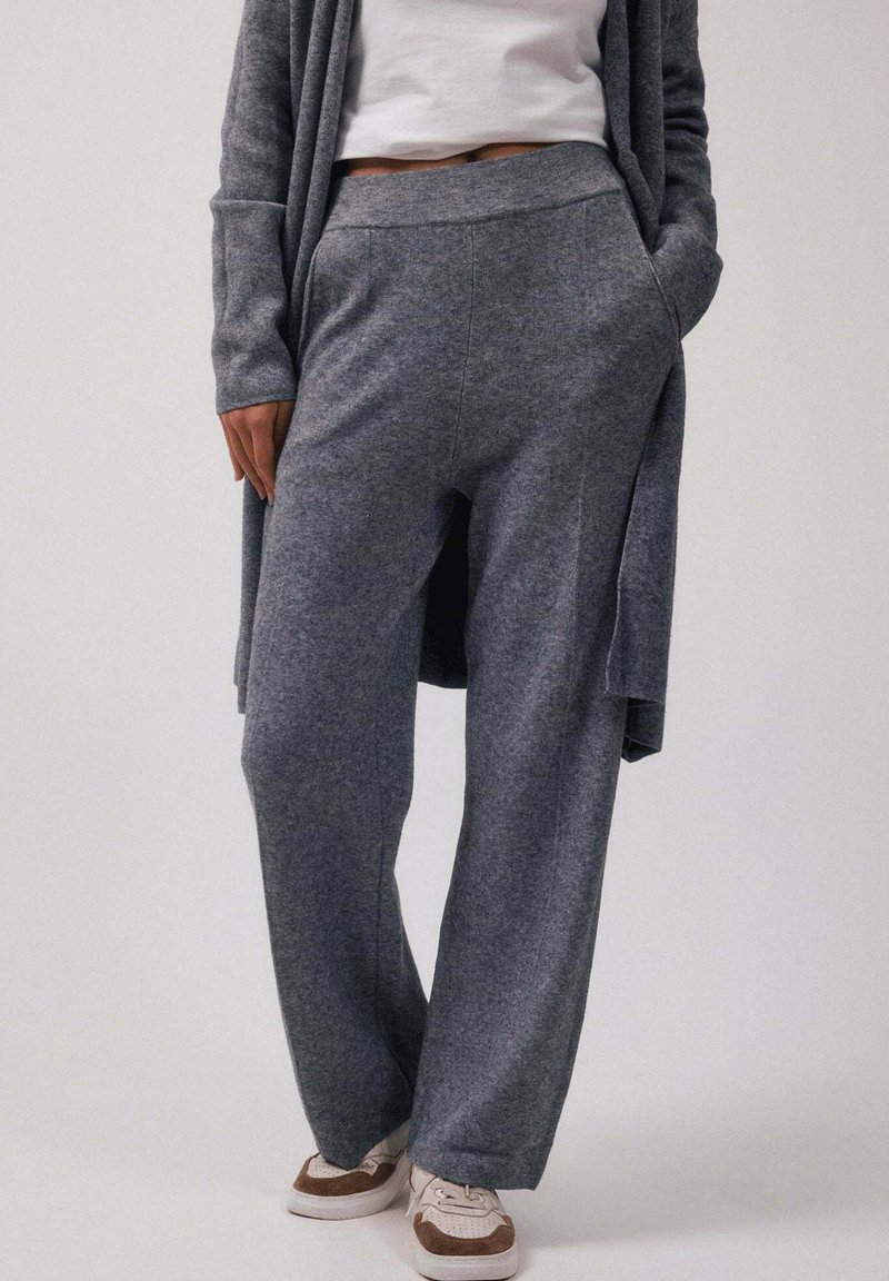 Personne portant un pantalon en maille gris ample et un cardigan assorti par-dessus un top court blanc, debout les mains dans les poches et des baskets blanches.