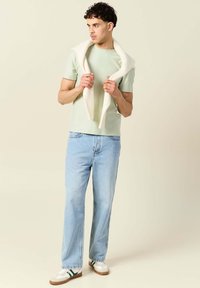 Ljusgrön t-shirt med kort ärm, blå rak ben jeans och vita sneakers med gröna detaljer; en krämfärgad stickad tröja hängande över axlarna.