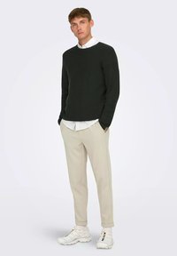 Maglione a maglia verde scuro con scollatura rotonda, pantaloni beige a sigaretta e sneaker bianche con dettagli colorati. Il modello sta in piedi con le mani in tasca.