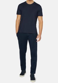 Man som bär en figurnära marinblå t-shirt med kort ärm, matchande smala byxor och marinblå sneakers med vita sulor, stående med en hand i fickan.