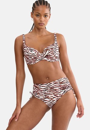 Kobieta z warkoczami, ubrana w bikini w zwierzęcy wzór zebry z wysokim stanem, stojąca na jasnym, gładkim tle.