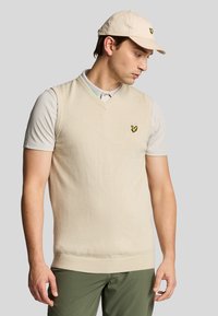 Maglione beige senza maniche con scollatura a V, tessuto testurizzato e un piccolo logo giallo sul petto, indossato sopra una polo grigia chiara.