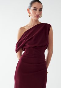 Vínová šaty na jedno rameno s drapovanou tkaninou, priliehajúcim korzetom a elegantnou siluetou. Má štruktúrovaný pás a hladkú textúru. Zlatučký náušnice ako doplnok.