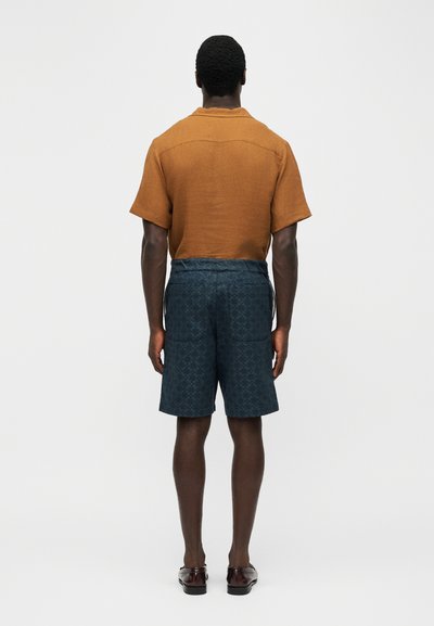 Άνδρας όρθιος, γυρισμένος προς τα πίσω, φορά κοντομάνικο πουκάμισο σε σκουριά απόχρωση, σκούρα σορτς με σχέδια και πίσω τσέπες, και καφέ παπούτσια.